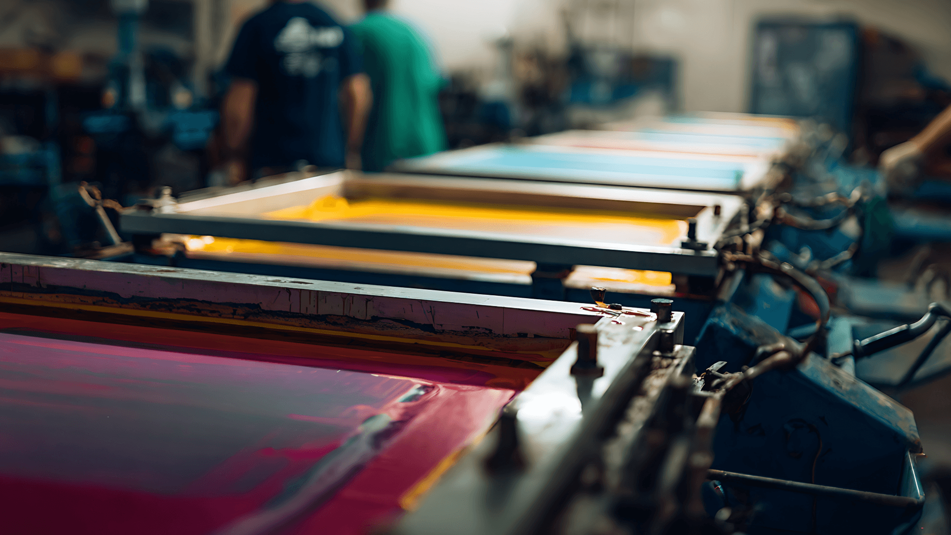 Vibrant pink, yellow, and blue ink inside screen printing frames in a blurred workshop setting.